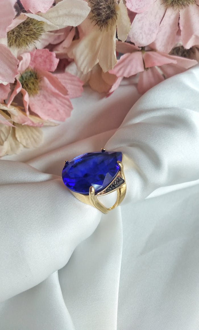 Close-up of a luxurious blue gemstone gold ring on white silk with delicate pink flowers.