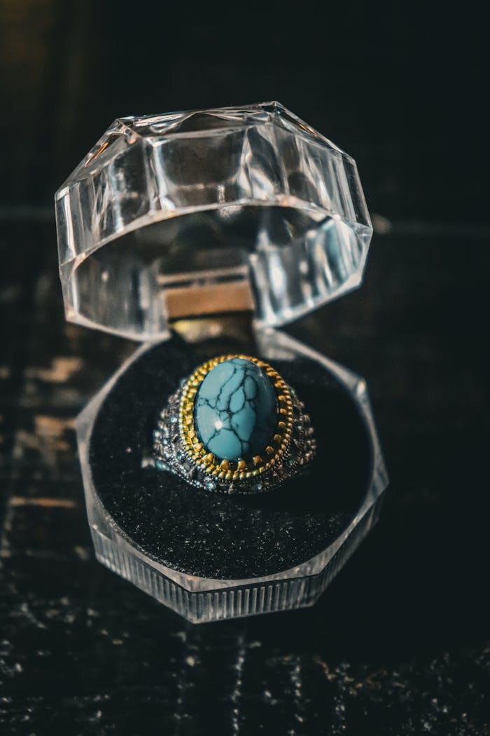 A beautifully detailed turquoise ring presented in a clear crystal box on a dark wooden surface.